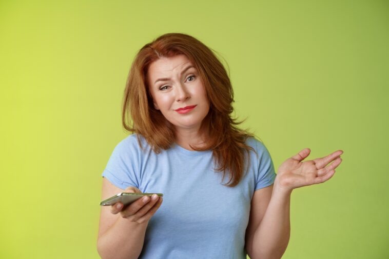 Woman with reddish-brown hair, wearing a blue t-shirt, holds a phone in one hand and shrugs with the other against a green background.