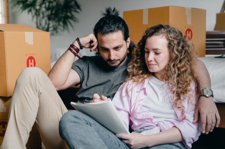 A couple sits on the floor surrounded by moving boxes, looking at a document together.
