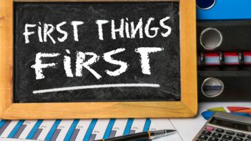 A chalkboard with the phrase "First Things First" is centered, surrounded by a wooden frame. Below, a financial chart, a pen, and a calculator are visible. Stacks of binders sit to the right.