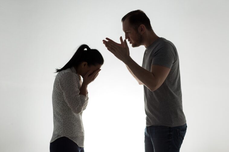 Silhouetted man gesturing emphatically at a woman who is covering her face with her hands.