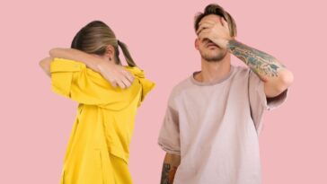 Two people against a solid pink background: a woman in a yellow shirt with her face covered by her arms, and a man in a light pink shirt with tattoos on his arm, covering his eyes with his hand.