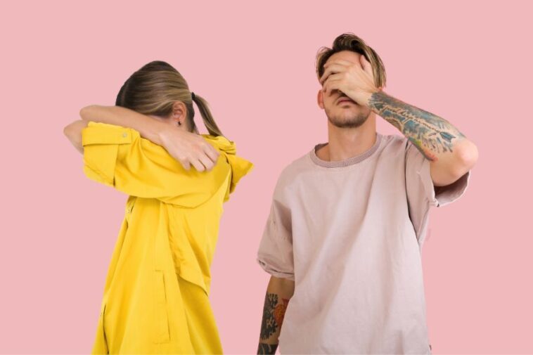 Two people against a solid pink background: a woman in a yellow shirt with her face covered by her arms, and a man in a light pink shirt with tattoos on his arm, covering his eyes with his hand.