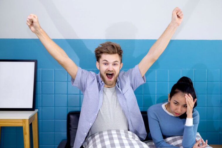 A man with arms raised in excitement sits next to a woman with her hand on her forehead, against a blue tiled wall.