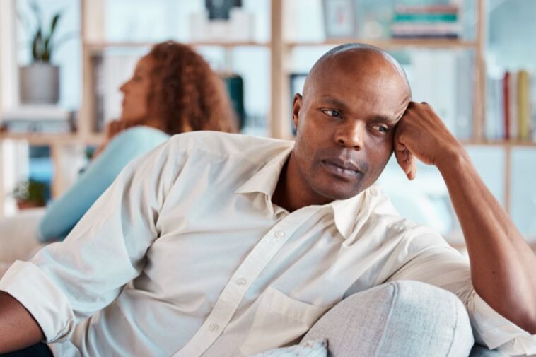 A man with a shaved head rests his face on his hand while sitting on a couch, with a woman in the background facing away from him.