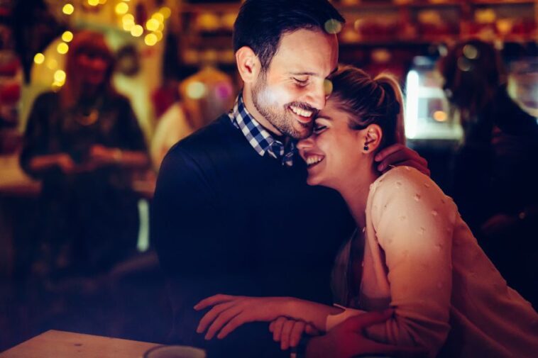 A smiling man and woman are embracing in a warmly lit, crowded indoor setting.