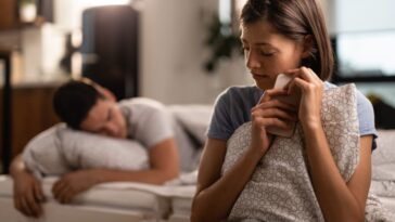 A woman in bed holds a phone and pillow, with a man asleep in the background.