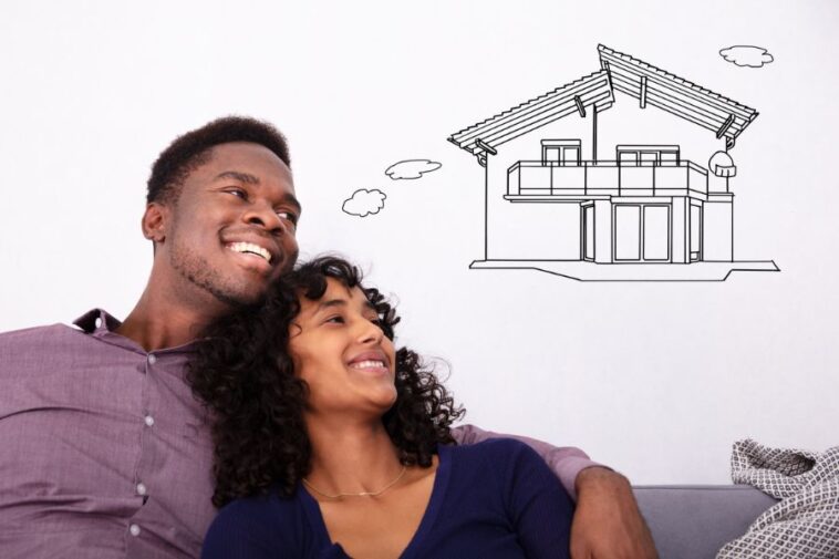 Smiling couple looking up at a drawing of a house on a white wall.