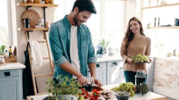 In a bright kitchen, a man in a denim shirt chops vegetables on a table with wine glasses, while a smiling woman holds a salad. Shelves and a ladder are visible in the background.