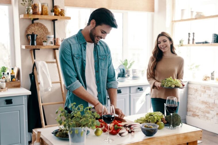 In a bright kitchen, a man in a denim shirt chops vegetables on a table with wine glasses, while a smiling woman holds a salad. Shelves and a ladder are visible in the background.