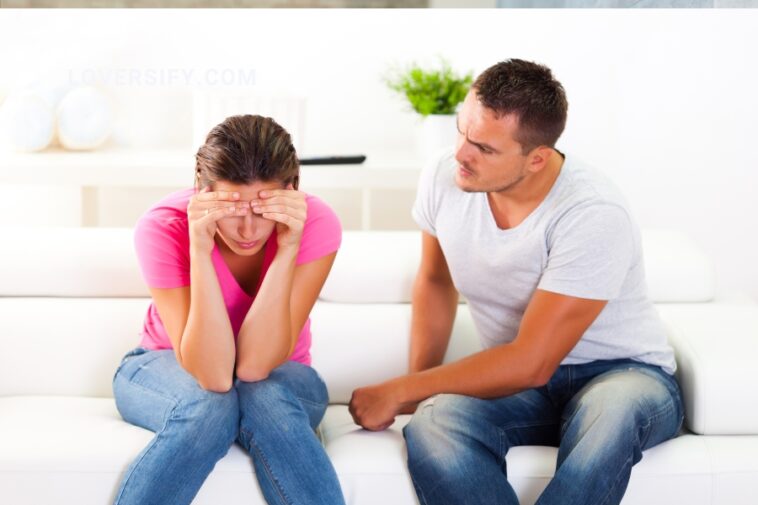A woman with her head in her hands sits on a white sofa next to a man who is looking at her with concern.