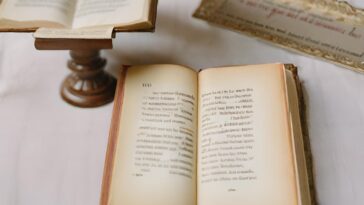 Open antique book with handwritten text, another book on a wooden stand, and a document with ornate edges on a white surface.