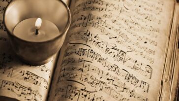 Open book of sheet music with a lit candle in a metal holder sitting on top.