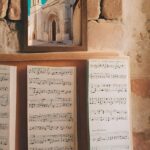 Sheets of music sit below a framed picture of a stone building. The background is a stone wall.