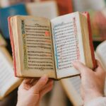 Hands hold open a small, old book with red edges and decorated borders, surrounded by other books.