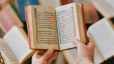 Hands hold open a small, old book with red edges and decorated borders, surrounded by other books.