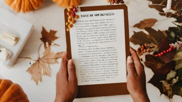Two hands hold a clipboard with text, surrounded by pumpkins, fall leaves, and decorative berries on a white surface.