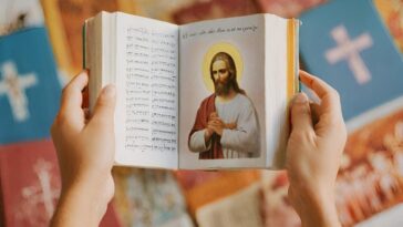 Hands hold open a book with a portrait of Jesus on the right page, text on the left, and other books with crosses on the covers are blurred in the background.