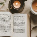 Open book of sheet music with two lit candles on a beige fabric surface.