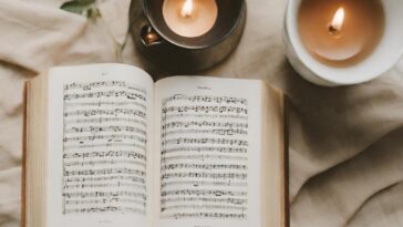 Open book of sheet music with two lit candles on a beige fabric surface.