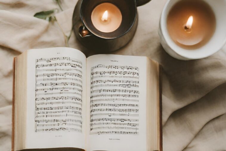 Open book of sheet music with two lit candles on a beige fabric surface.