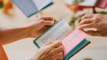 Two people are holding open small books with text inside, one with a pink page featuring a cross. Other similar books are visible in the background.