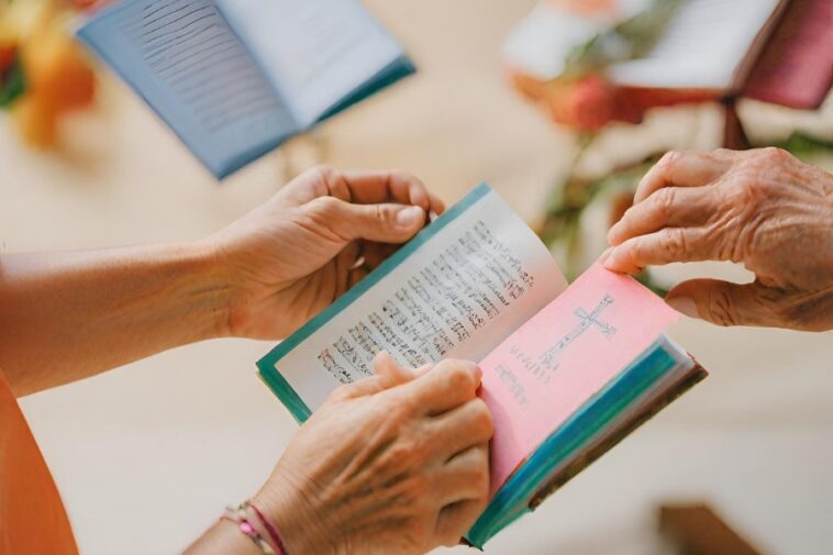 Two people are holding open small books with text inside, one with a pink page featuring a cross. Other similar books are visible in the background.