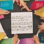 A sheet of music sits in the center of a dark cloth, surrounded by colorful papers with musical notations and hands reaching in.