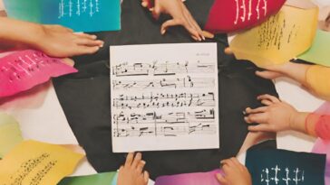 A sheet of music sits in the center of a dark cloth, surrounded by colorful papers with musical notations and hands reaching in.