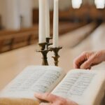 Close-up of a person reading an open book in a church, with a brass candle holder and white candles nearby. Rows of pews are visible in the background.