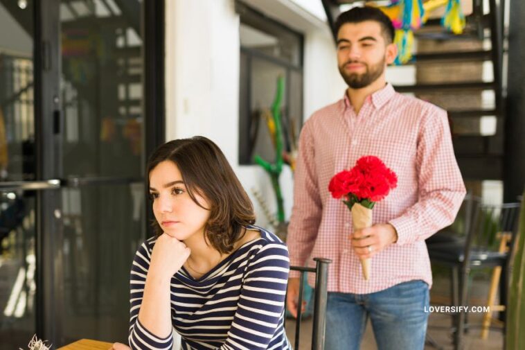 A woman sits with a bored or upset expression at a table while a man stands behind her holding a bouquet of red flowers.