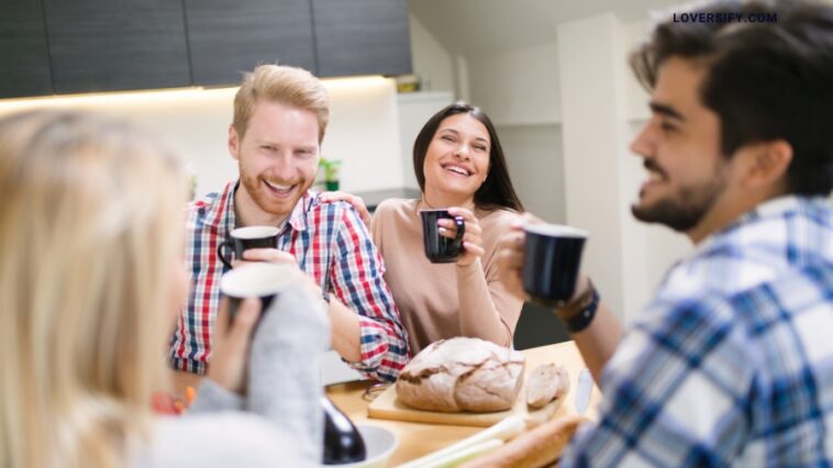 Four friends smiling and drinking coffee around a kitchen table, with bread and vegetables in front of them.