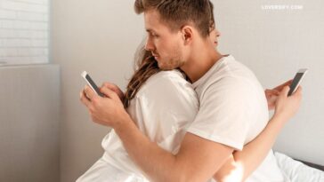 A man and a woman are hugging while looking at their phones. They are both wearing white shirts.