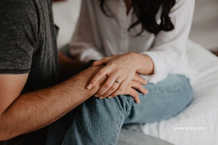 A woman in jeans sits on a white cushion holding a man's arm, showing off a diamond ring.
