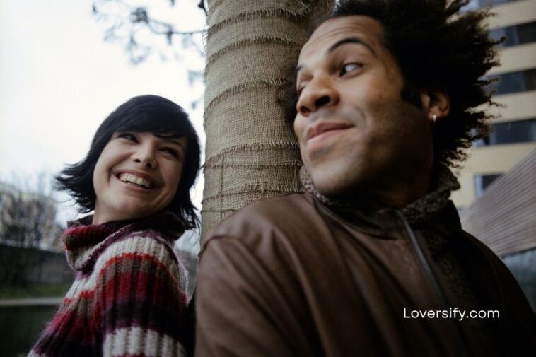 A smiling woman in a striped sweater leans against a tree next to a man in a brown jacket who looks away, against an urban background.