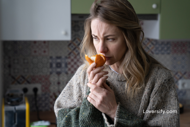 Woman in a kitchen holds a peeled orange to her nose with a troubled expression.