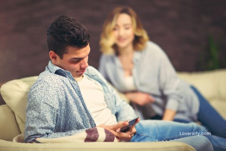 A young man sits on a cream couch, focused on a smartphone, while a woman in the background smiles and relaxes on the same couch.