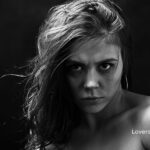 Black and white portrait of a woman with intense, direct gaze, partially bare shoulders, and dark smudges around eyes. Her hair is windswept.
