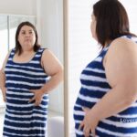 Woman in a blue and white striped dress is looking at herself in a mirror, touching her abdomen.
