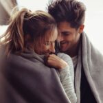 A young couple, wrapped in a gray blanket, are smiling and looking at each other affectionately outdoors.