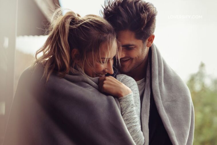 A young couple, wrapped in a gray blanket, are smiling and looking at each other affectionately outdoors.