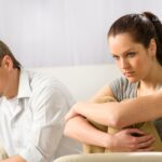 A man and a woman sit apart on a sofa, both looking down with somber expressions.