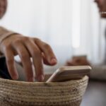 A hand reaches into a woven basket containing several smartphones, with two people blurred in the background.