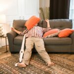 A person in a plaid shirt and tan pants is lying on the floor in front of a couch with an orange pillow covering their face. A vacuum cleaner is attached to their leg.