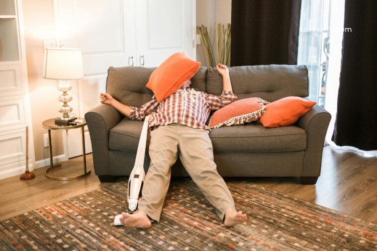 A person in a plaid shirt and tan pants is lying on the floor in front of a couch with an orange pillow covering their face. A vacuum cleaner is attached to their leg.