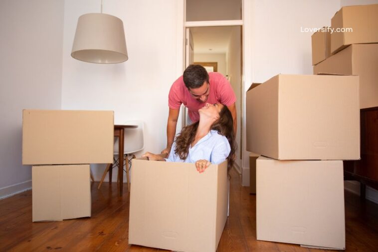 A man in a pink shirt leans over a woman sitting in a large cardboard box, both smiling at each other, surrounded by moving boxes in a room.