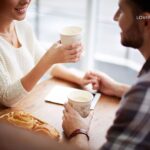 A man and woman hold coffee cups while sitting at a table with pastries.