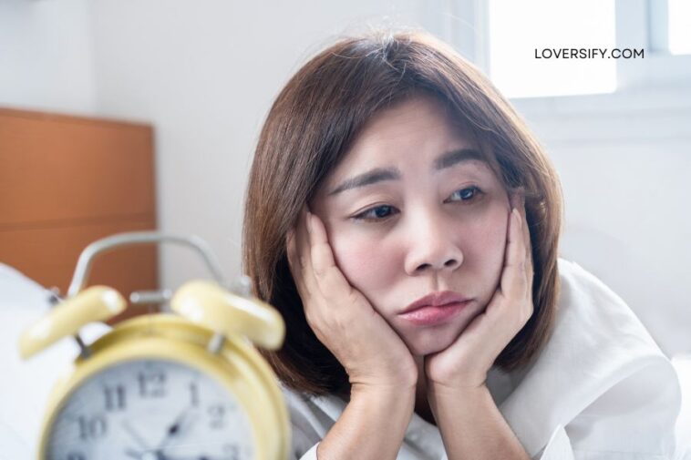 A woman with her hands on her face looks dejectedly at a yellow alarm clock in the foreground.