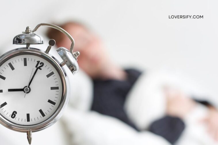 Close-up of a silver alarm clock in the foreground, with a blurred person in bed in the background.