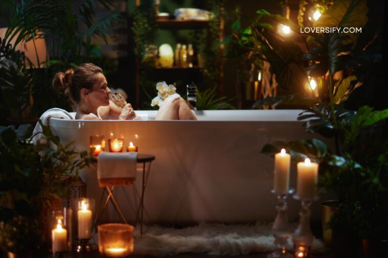A woman reclines in a bathtub surrounded by lush greenery and illuminated by warm candlelight.
