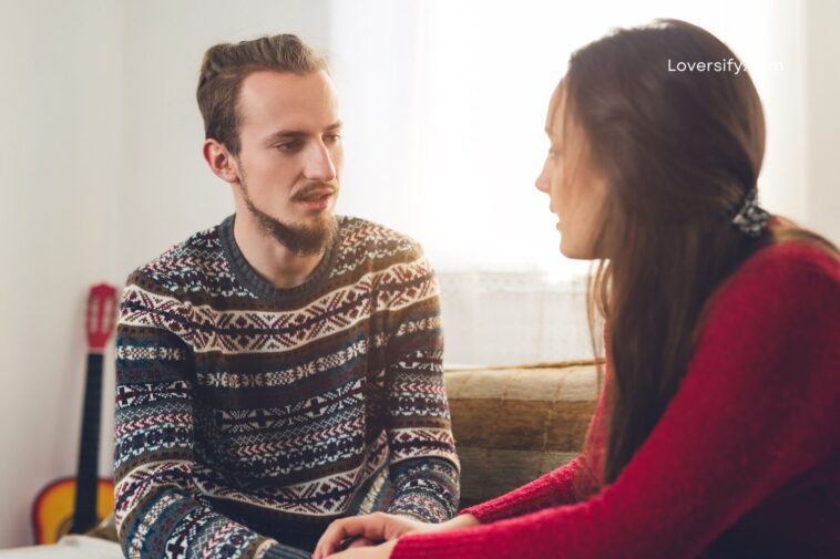 A man with a beard and a patterned sweater speaks to a woman in a red sweater, with a guitar in the background.